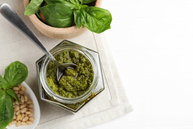 Photo of Tasty pesto sauce in jar, spoon, pine nuts and basil on white table, top view. Space for text