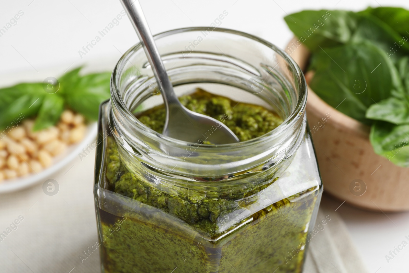 Photo of Tasty pesto sauce in glass jar, spoon, pine nuts and basil on table, closeup