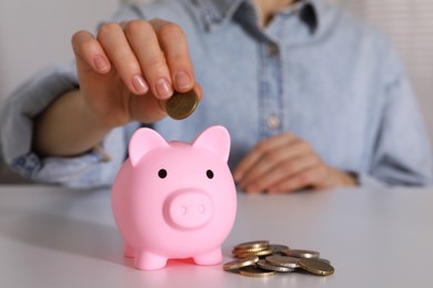 Woman putting coin into pink piggy bank at white table, closeup. Space for text