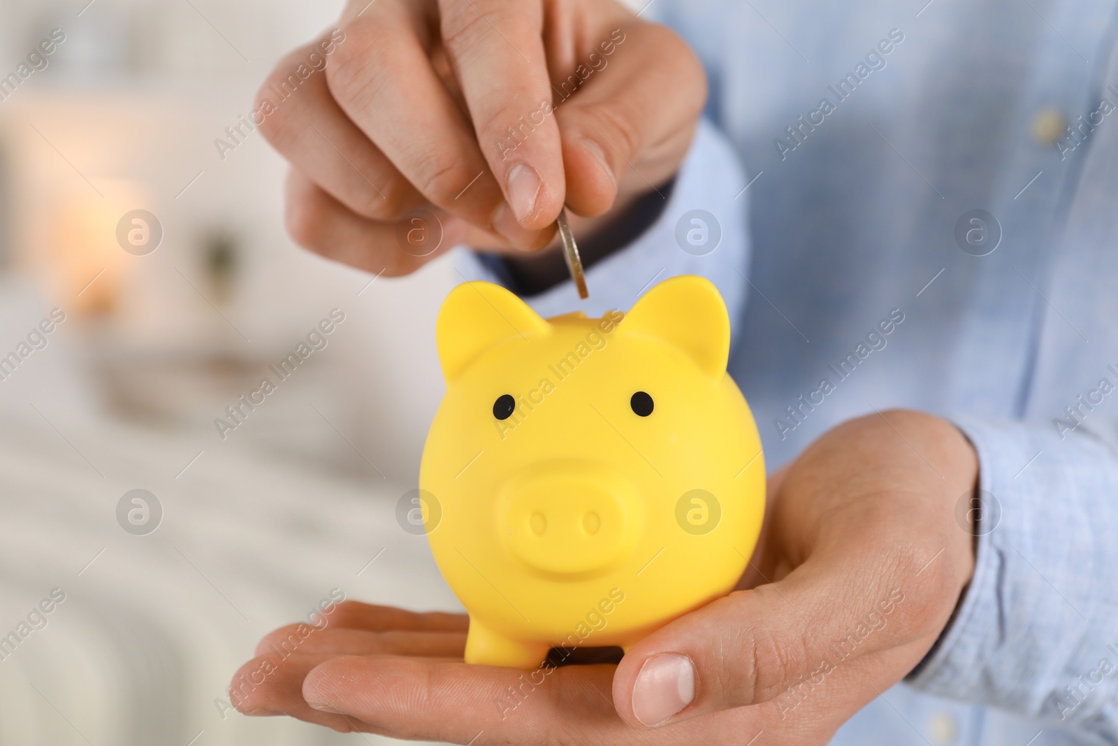 Photo of Man putting coin into yellow piggy bank indoors, closeup