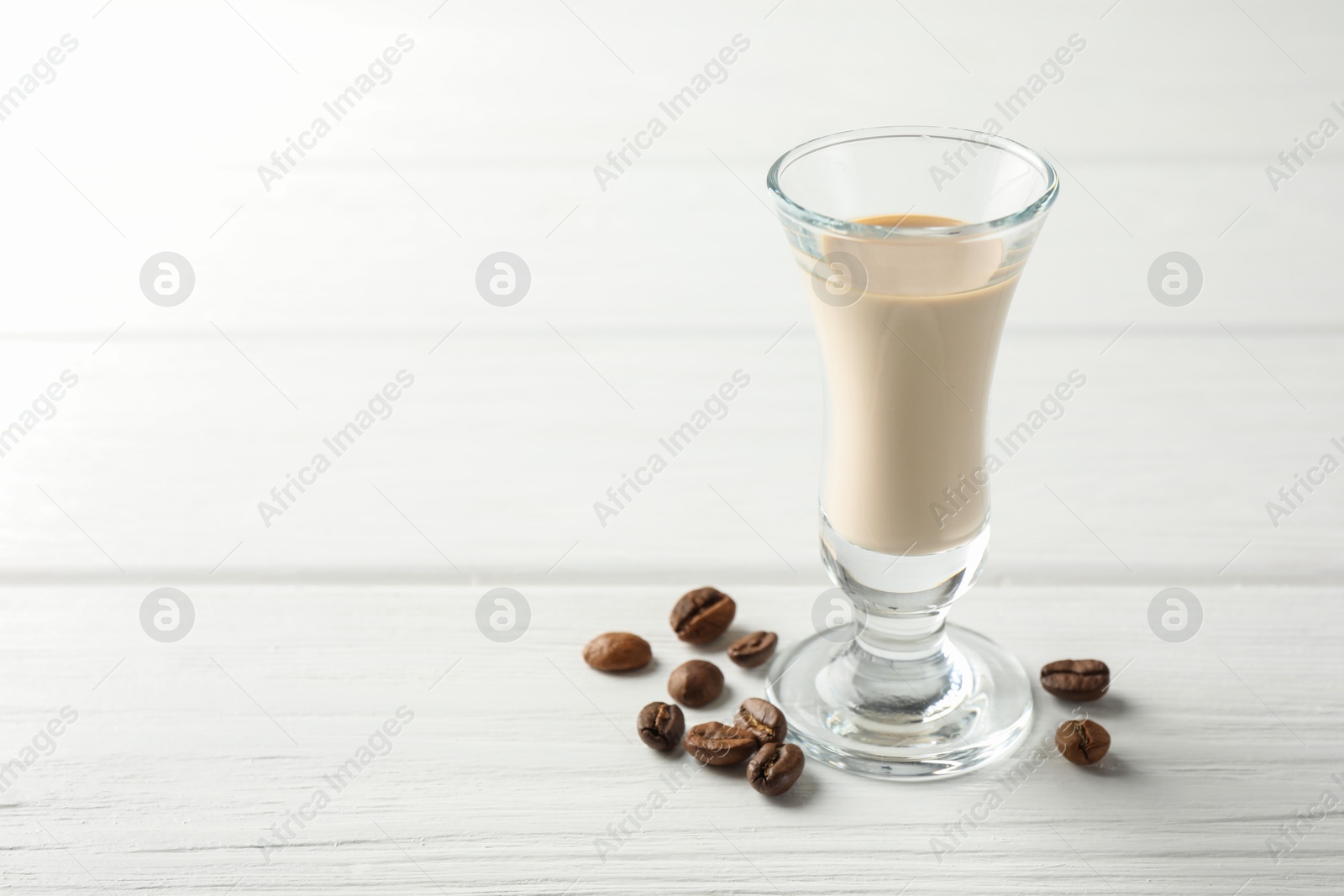 Photo of Coffee cream liqueur in glass and beans on white wooden table, space for text