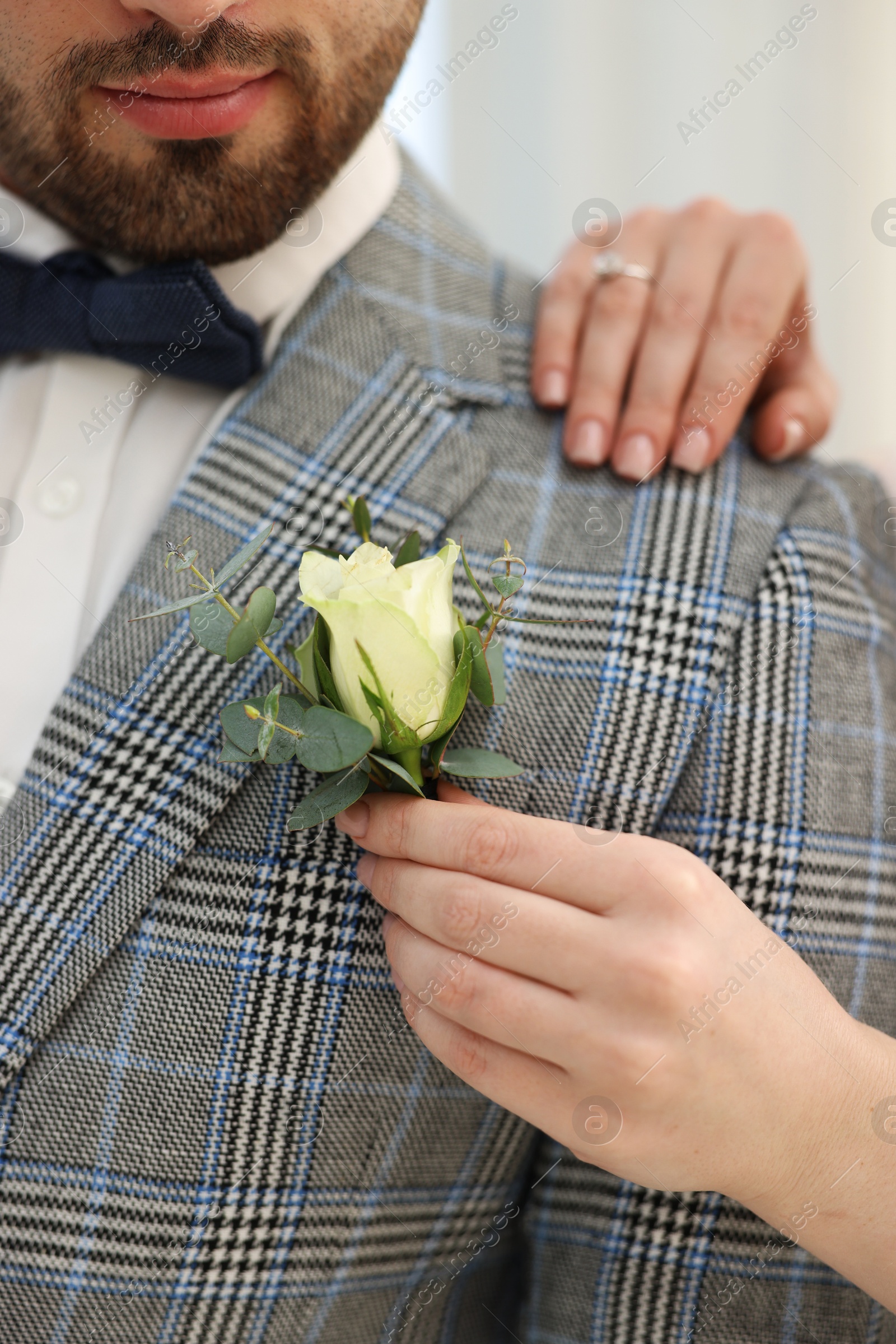 Photo of Bride putting boutonniere on her groom's jacket against light background, closeup. Wedding accessory