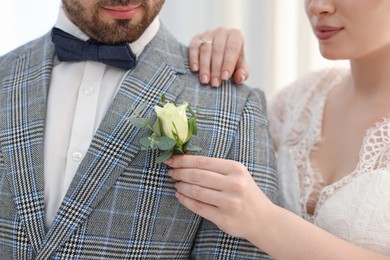 Photo of Bride putting boutonniere on her groom's jacket against light background, closeup. Wedding accessory