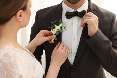 Photo of Bride putting boutonniere on her groom's jacket against light background, closeup. Wedding accessory