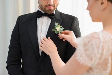 Bride putting boutonniere on her groom's jacket against light background, closeup. Wedding accessory