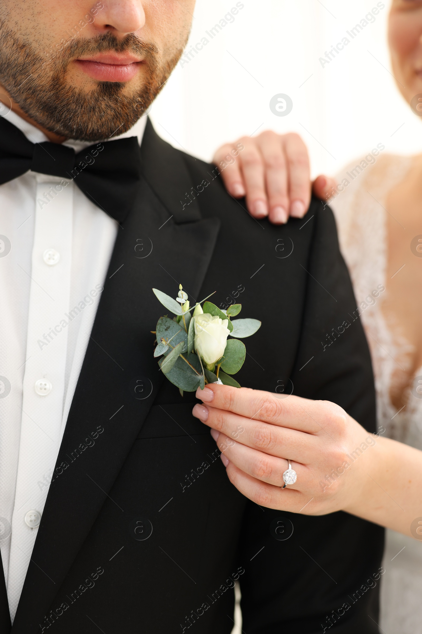 Photo of Bride putting boutonniere on her groom's jacket against light background, closeup. Wedding accessory