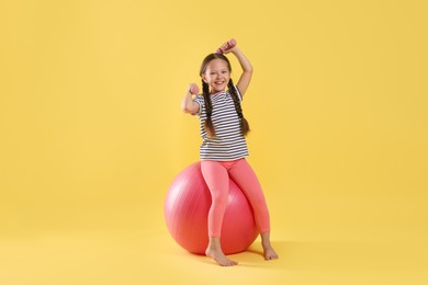 Photo of Cute little girl with dumbbells and fit ball on yellow background