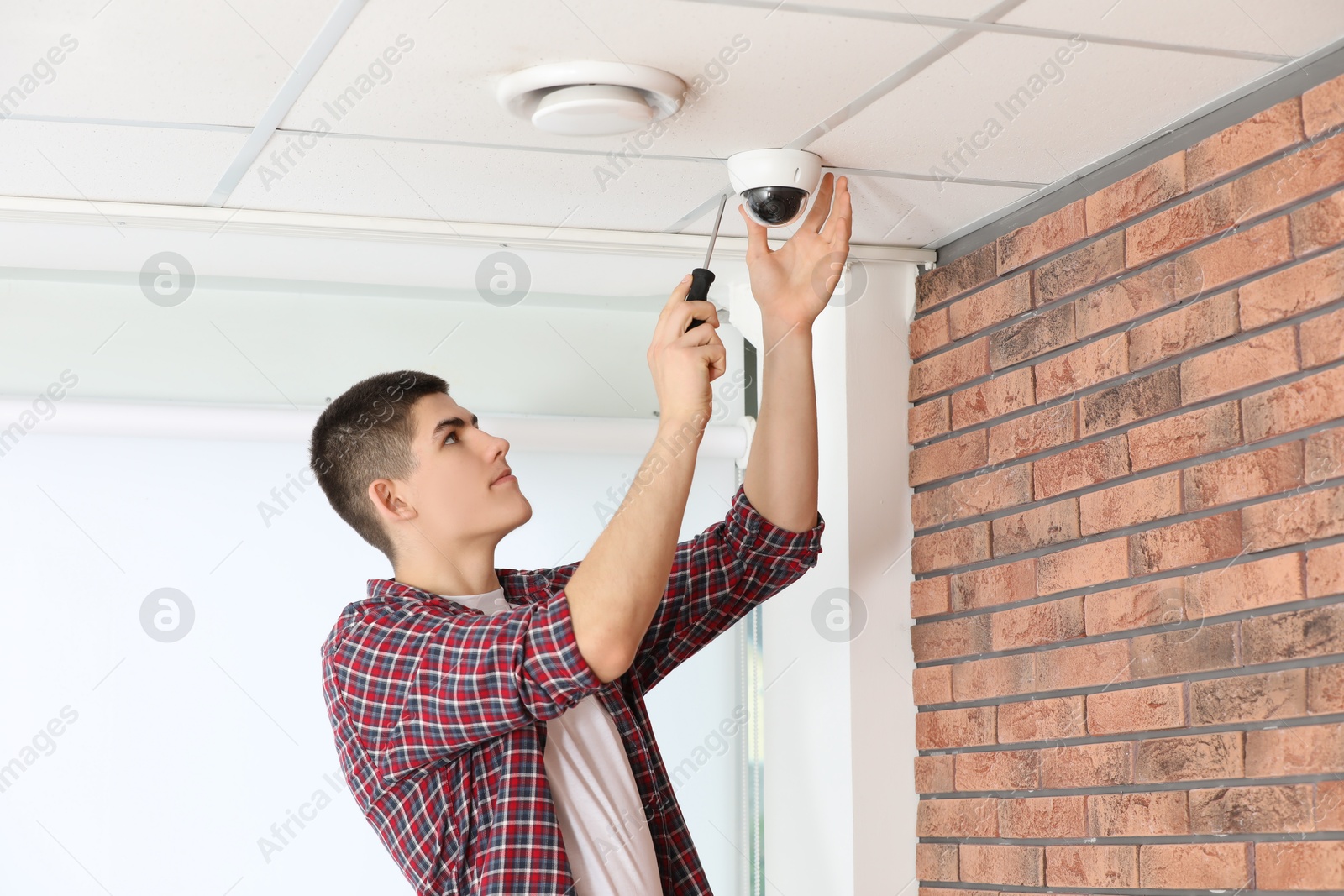 Photo of Technician with screwdriver installing CCTV camera on ceiling indoors