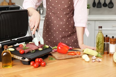 Photo of Woman cooking different products with electric grill at wooden table in kitchen, closeup