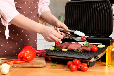 Woman cooking different products with electric grill at wooden table in kitchen, closeup