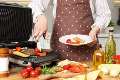 Photo of Woman cooking different products with electric grill at wooden table in kitchen, closeup