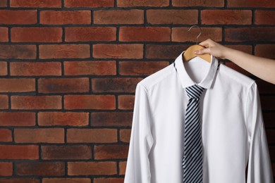 Woman holding hanger with shirt and necktie near red brick wall, space for text