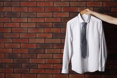 Woman holding hanger with shirt and necktie near red brick wall, space for text