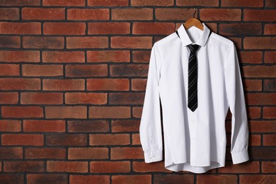 Hanger with white shirt and striped necktie on red brick wall, space for text