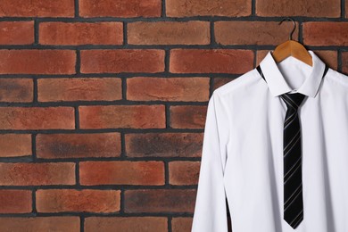 Hanger with white shirt and striped necktie on red brick wall, space for text