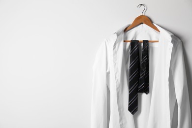 Photo of Hanger with white shirt and striped necktie on light wall, space for text