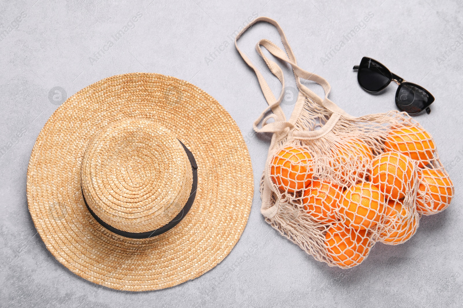 Photo of String bag with oranges, sunglasses and straw hat on grey table, top view