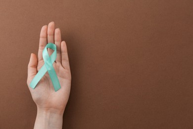 Photo of Woman holding turquoise awareness ribbon on brown background, top view. Space for text