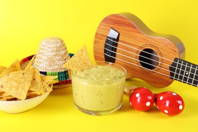 Photo of Delicious guacamole with nachos chips, Mexican sombrero hat, ukulele and maracas on yellow background