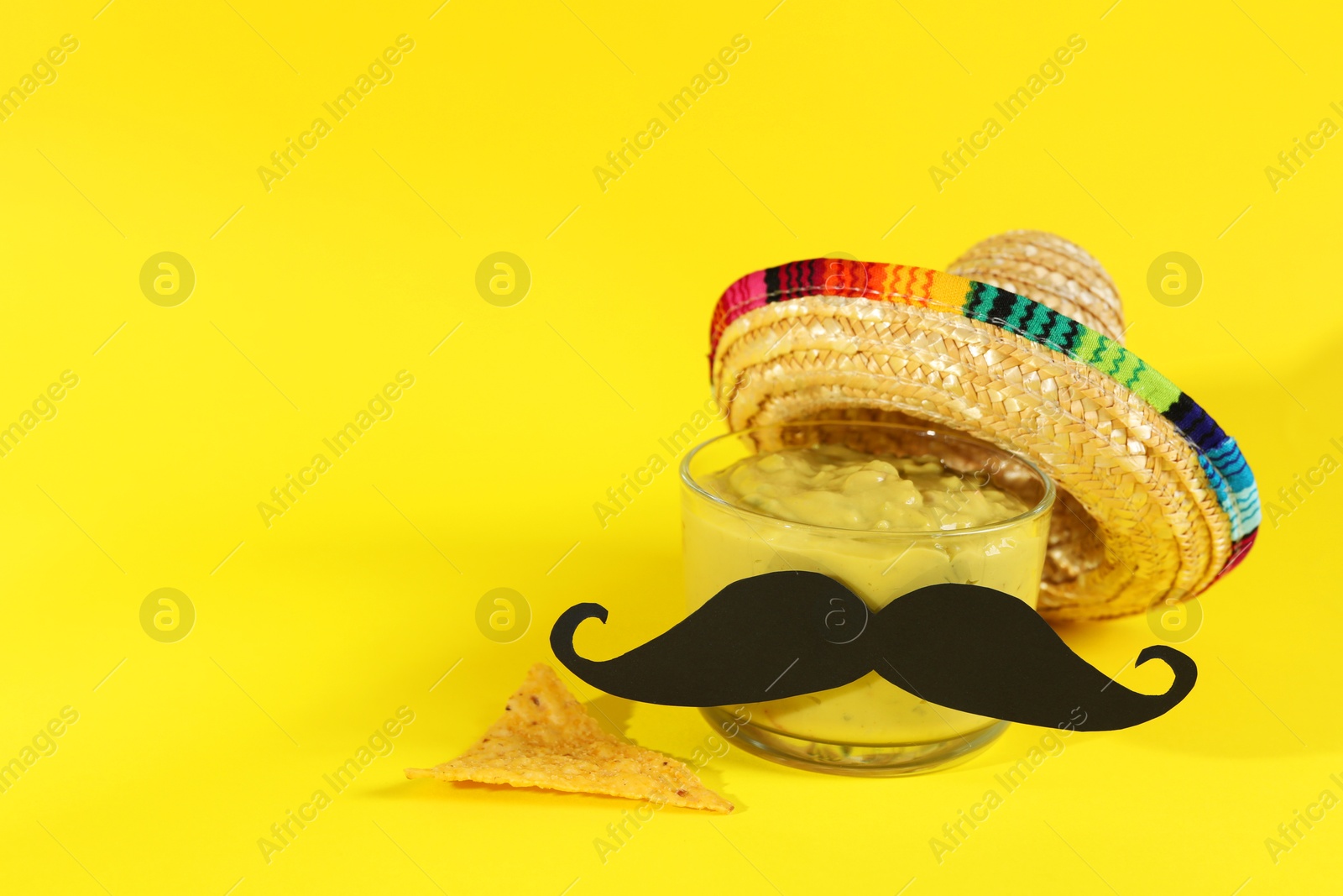 Photo of Delicious guacamole, nachos chip, Mexican sombrero hat and fake mustache on yellow background. Space for text