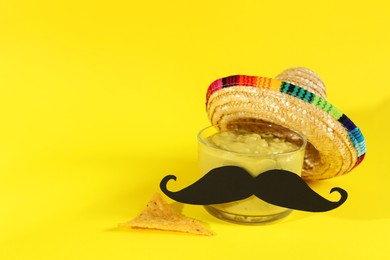 Photo of Delicious guacamole, nachos chip, Mexican sombrero hat and fake mustache on yellow background. Space for text