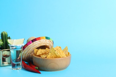 Photo of Mexican sombrero hat, chili peppers, nachos chips and tequila on light blue background. Space for text