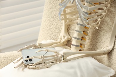 Photo of Waiting concept. Human skeleton with smartphone sitting in armchair indoors, closeup