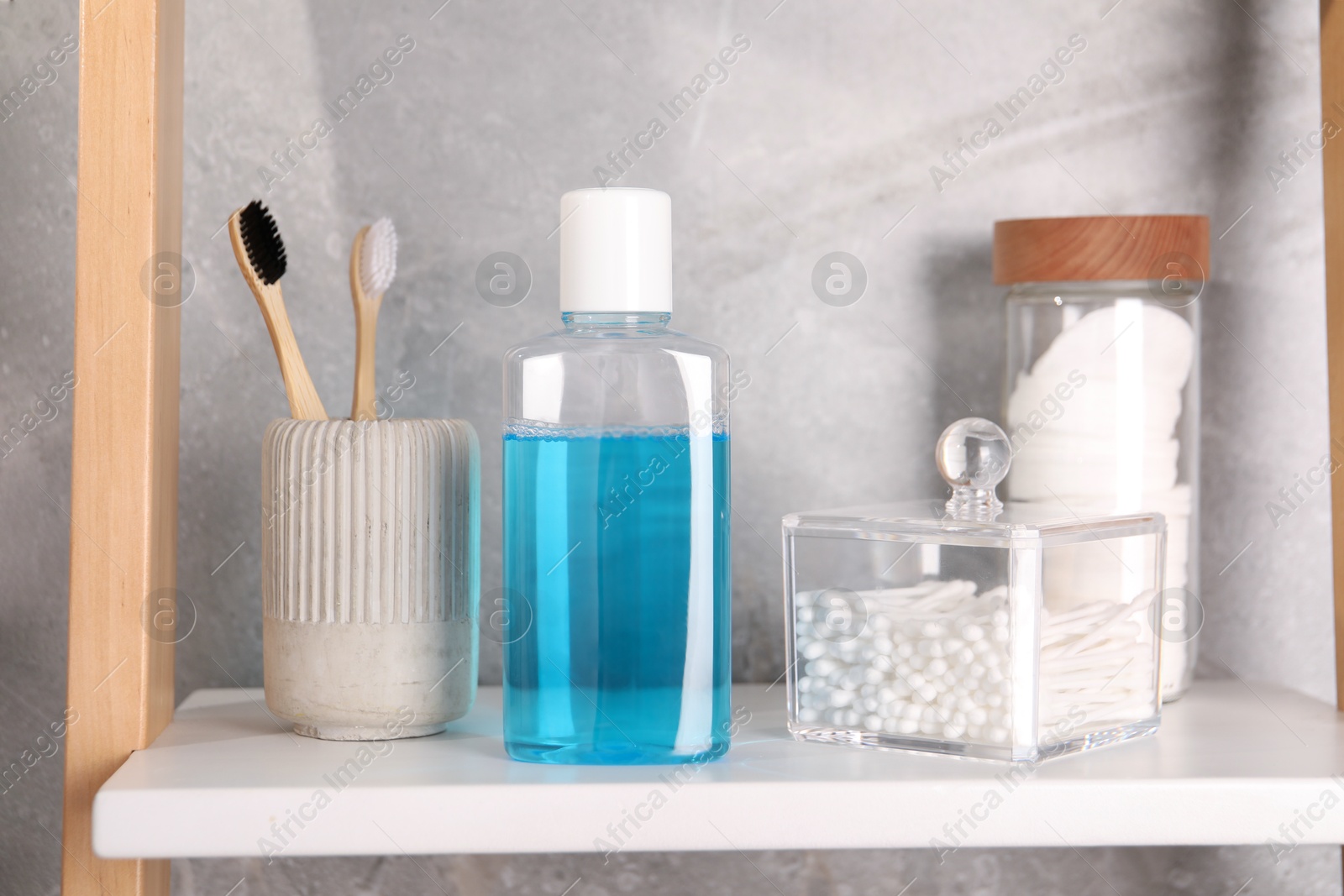 Photo of Bottle of mouthwash, toothbrushes, cotton buds and pads on white shelf in bathroom