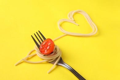 Heart made of tasty spaghetti, fork and tomato on yellow background, closeup