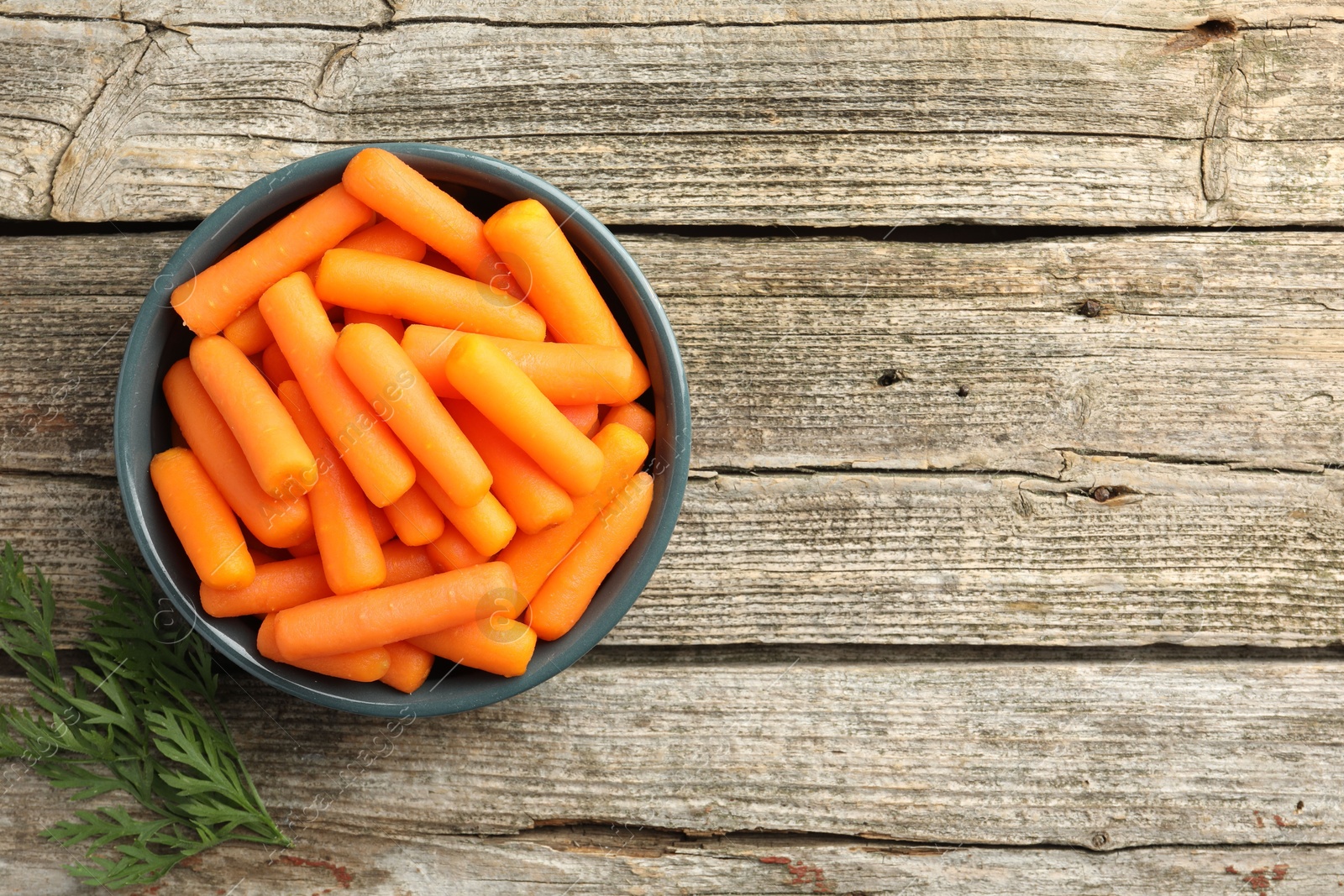 Photo of Baby carrots in bowl and green leaf on wooden table, top view. Space for text