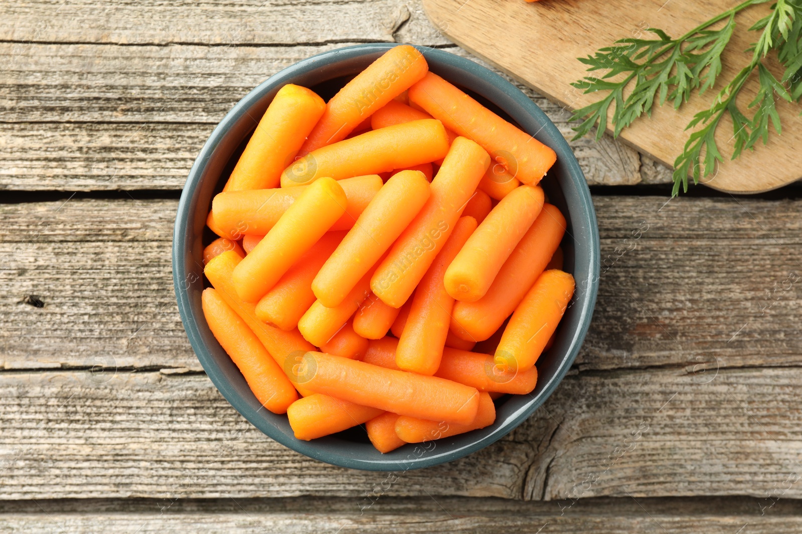 Photo of Baby carrots in bowl and green leaves on wooden table, top view