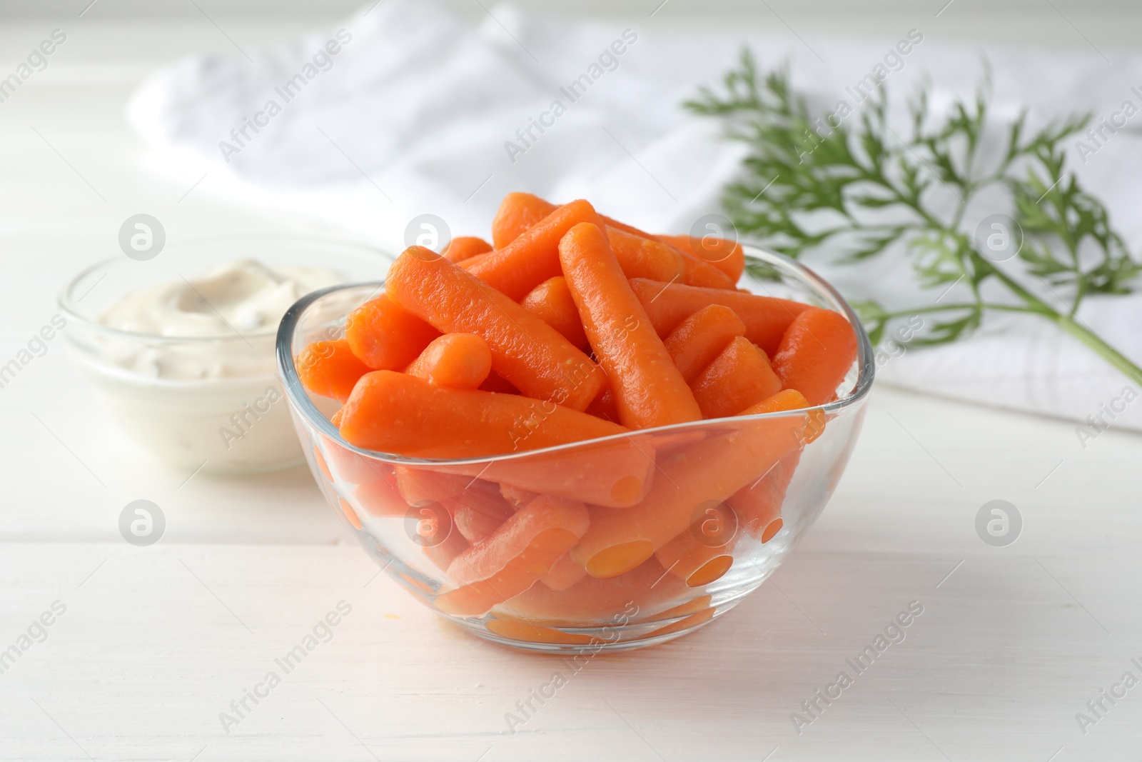 Photo of Baby carrots in glass bowl, sauce and green leaf on white wooden table