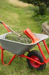 Wheelbarrow with mown grass, rubber boots and pitchfork outdoors