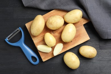 Photo of Fresh raw potatoes and peeler on black wooden table, flat lay