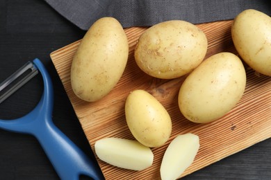 Fresh raw potatoes and peeler on black wooden table, flat lay