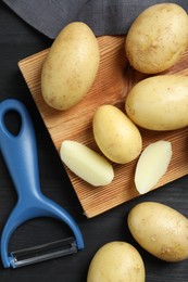 Fresh raw potatoes and peeler on black wooden table, flat lay