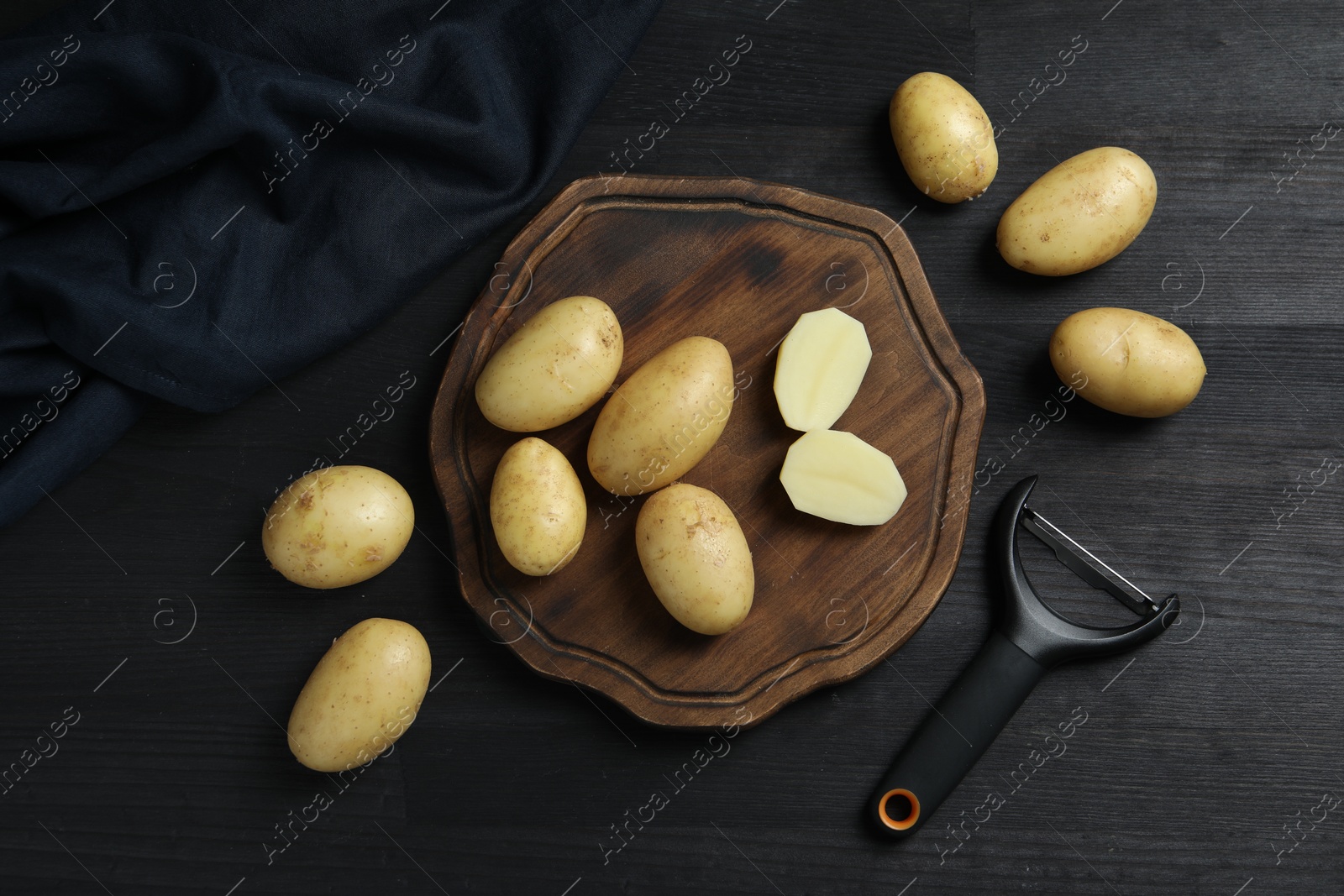 Photo of Fresh raw potatoes and peeler on black wooden table, flat lay