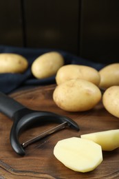 Fresh raw potatoes and peeler on cutting board against black background, closeup