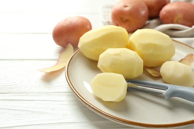 Fresh raw potatoes and peeler on white wooden table, closeup. Space for text