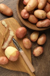 Photo of Fresh raw potatoes, peels and peeler on wooden table, flat lay