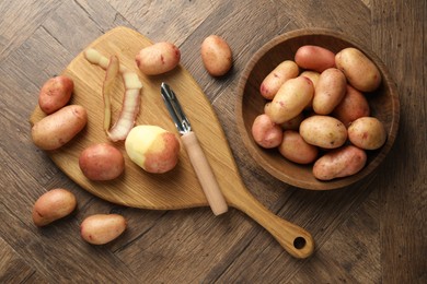 Fresh raw potatoes, peels and peeler on wooden table, flat lay