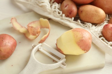 Photo of Fresh raw potatoes, peels and peeler on white table, closeup