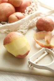 Fresh raw potatoes, peels and peeler on white wooden table