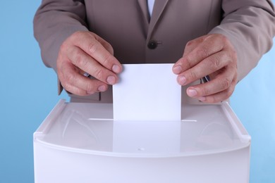 Man putting his vote into ballot box against light blue background, closeup