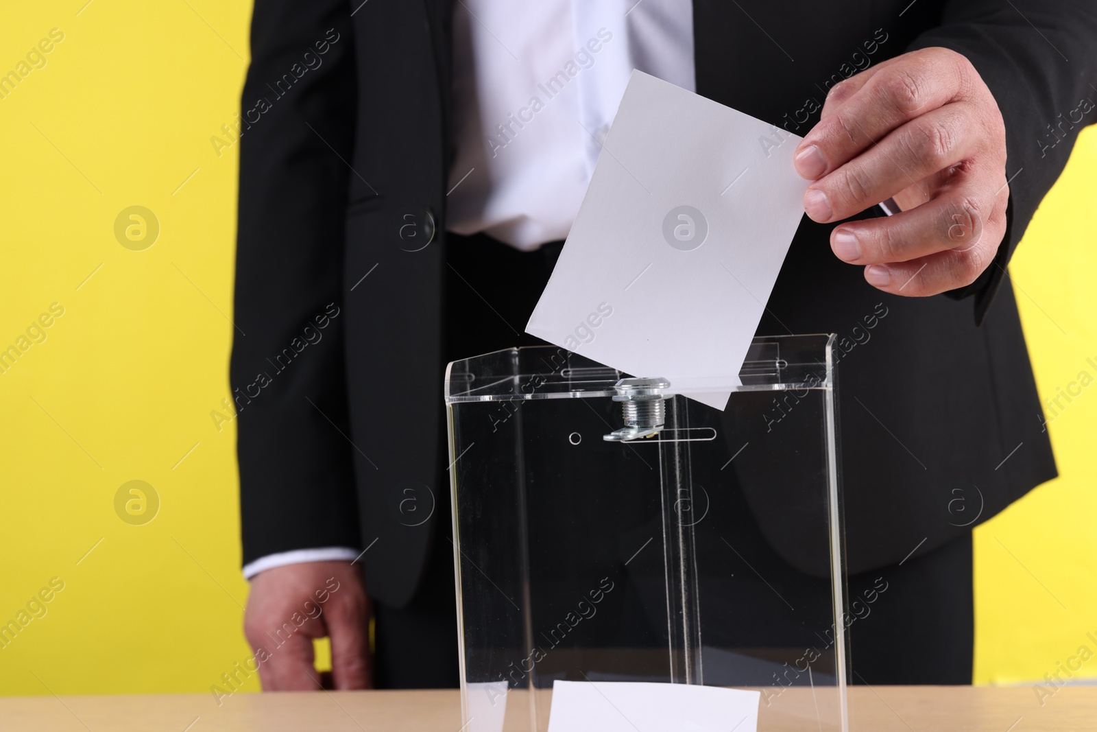 Photo of Man putting his vote into ballot box at table against yellow background, closeup