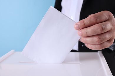 Man putting his vote into ballot box against light blue background, closeup