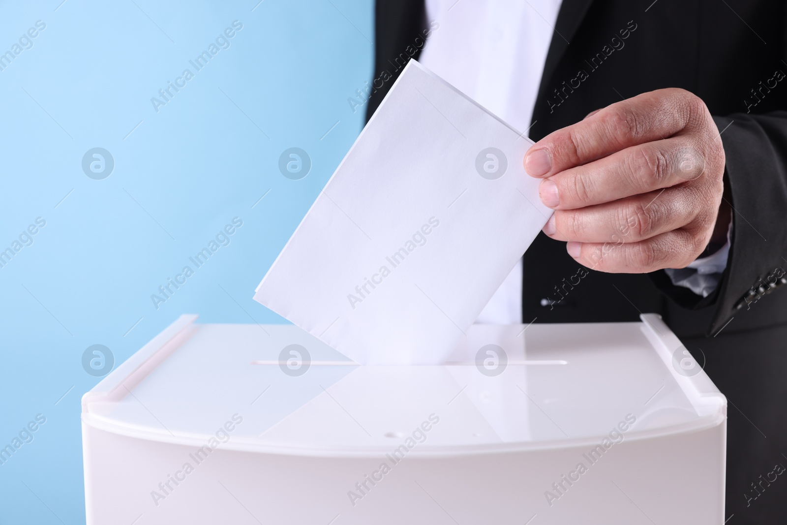 Photo of Man putting his vote into ballot box against light blue background, closeup