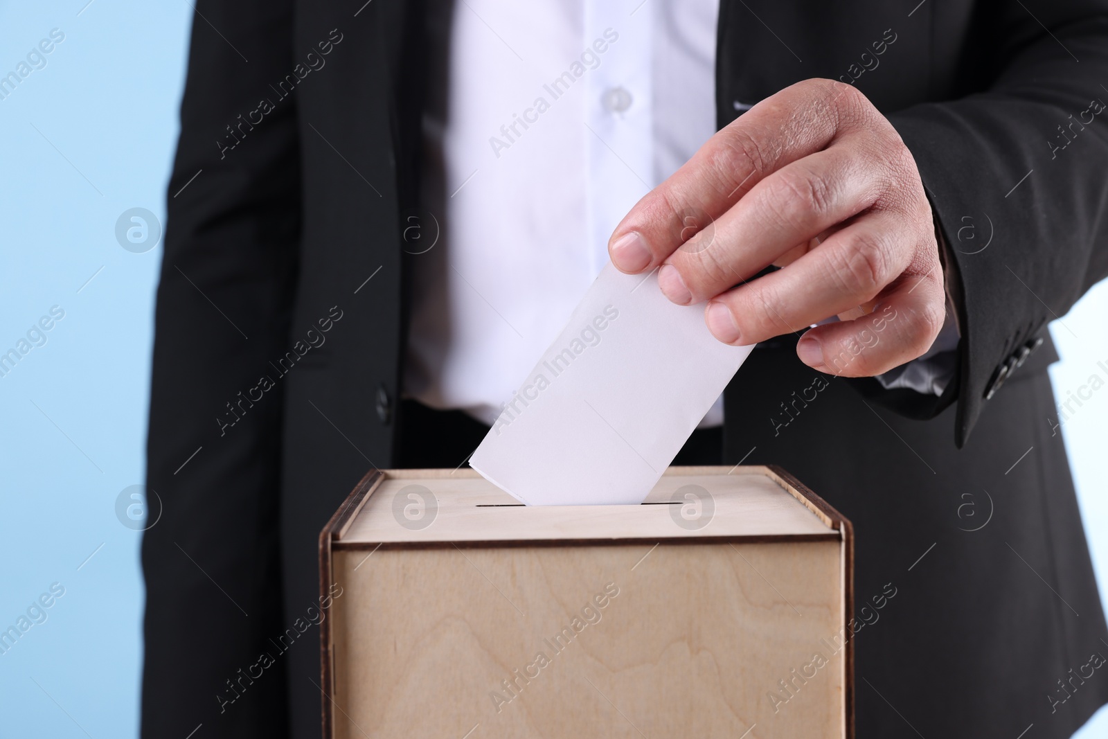 Photo of Man putting his vote into ballot box against light blue background, closeup