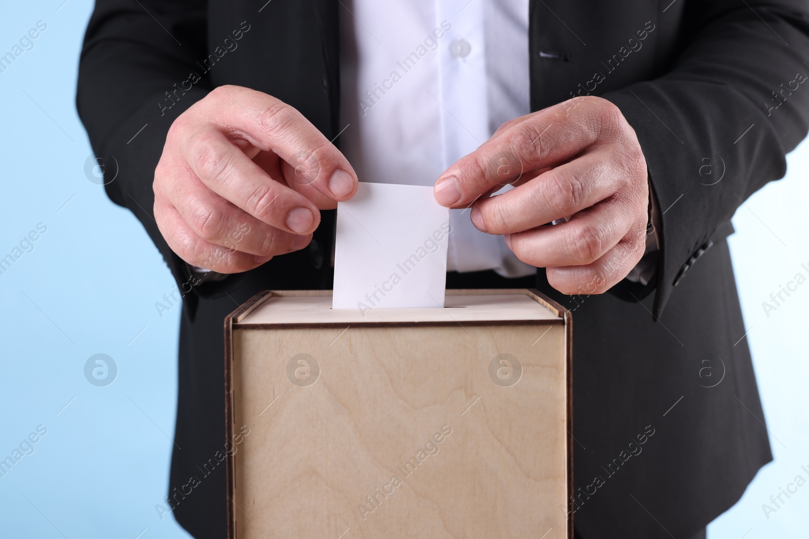 Photo of Man putting his vote into ballot box against light blue background, closeup