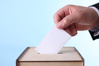 Photo of Man putting his vote into ballot box against light blue background, closeup